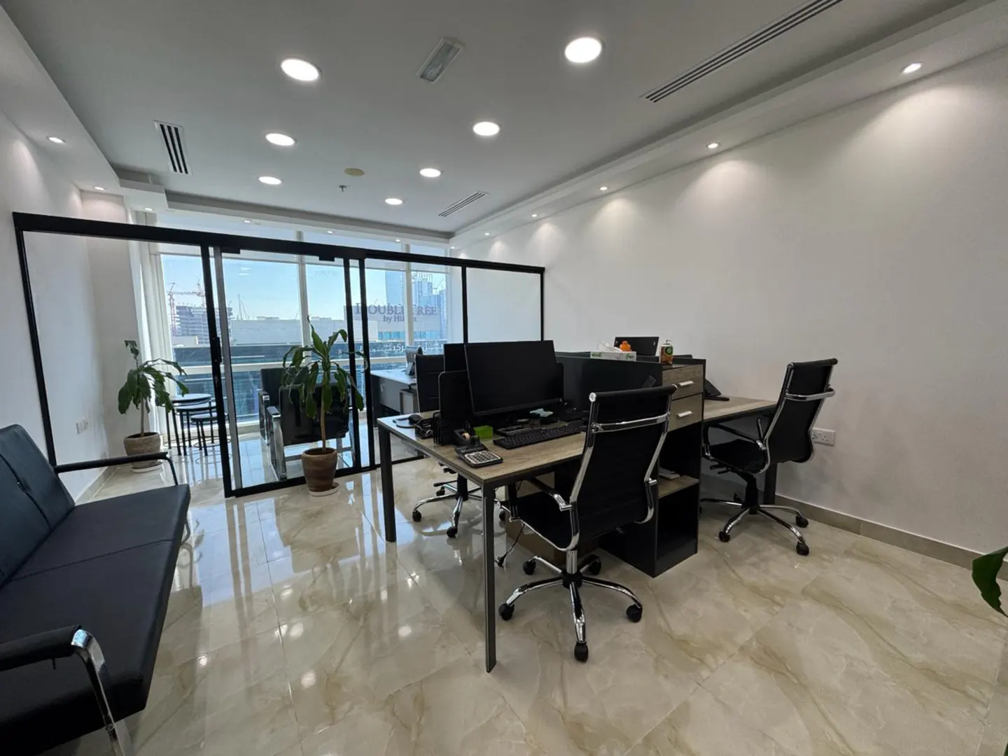 Modern office with two desks, black chairs, a sofa, potted plants, and large windows with a city view.