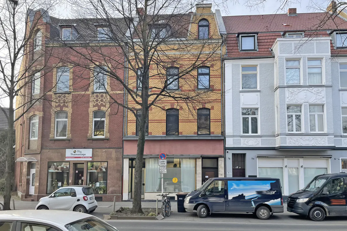 Street view of three brick buildings with storefronts, parked cars, and a bare tree in front.