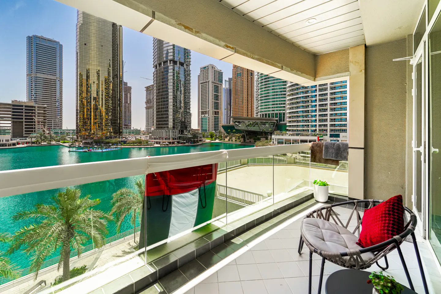 Balcony view of Dubai Marina with skyscrapers, turquoise water, and palm trees. A chair with a red pillow sits on the tiled floor.