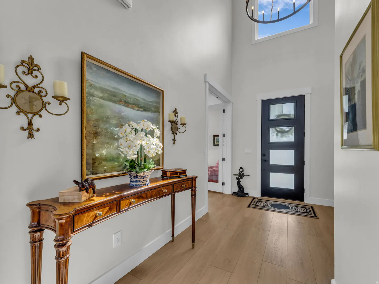 Foyer with a wood console table, white orchids, and a landscape painting. A black front door and chandelier are visible.