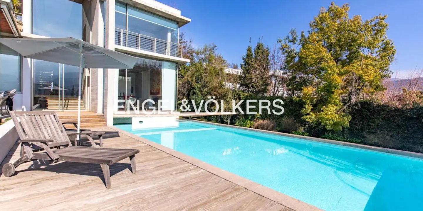 Modern home with a pool. Wooden deck with lounge chairs and umbrella. Engel & Völkers logo in the center. Trees and blue sky in the background.