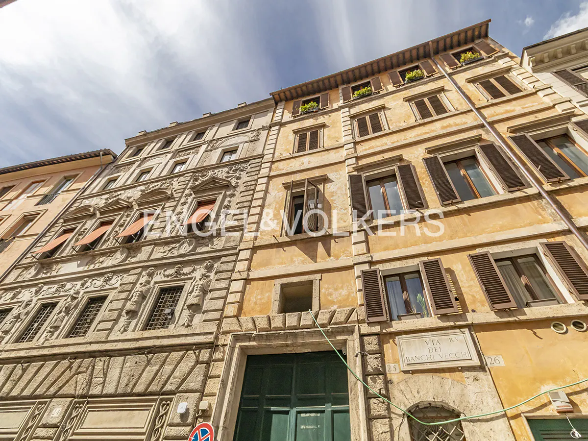 Exterior view of a multi-story building with stone facade and dark green doors. Windows have brown shutters. A street sign reads "Via dei Banchi Vecchi".