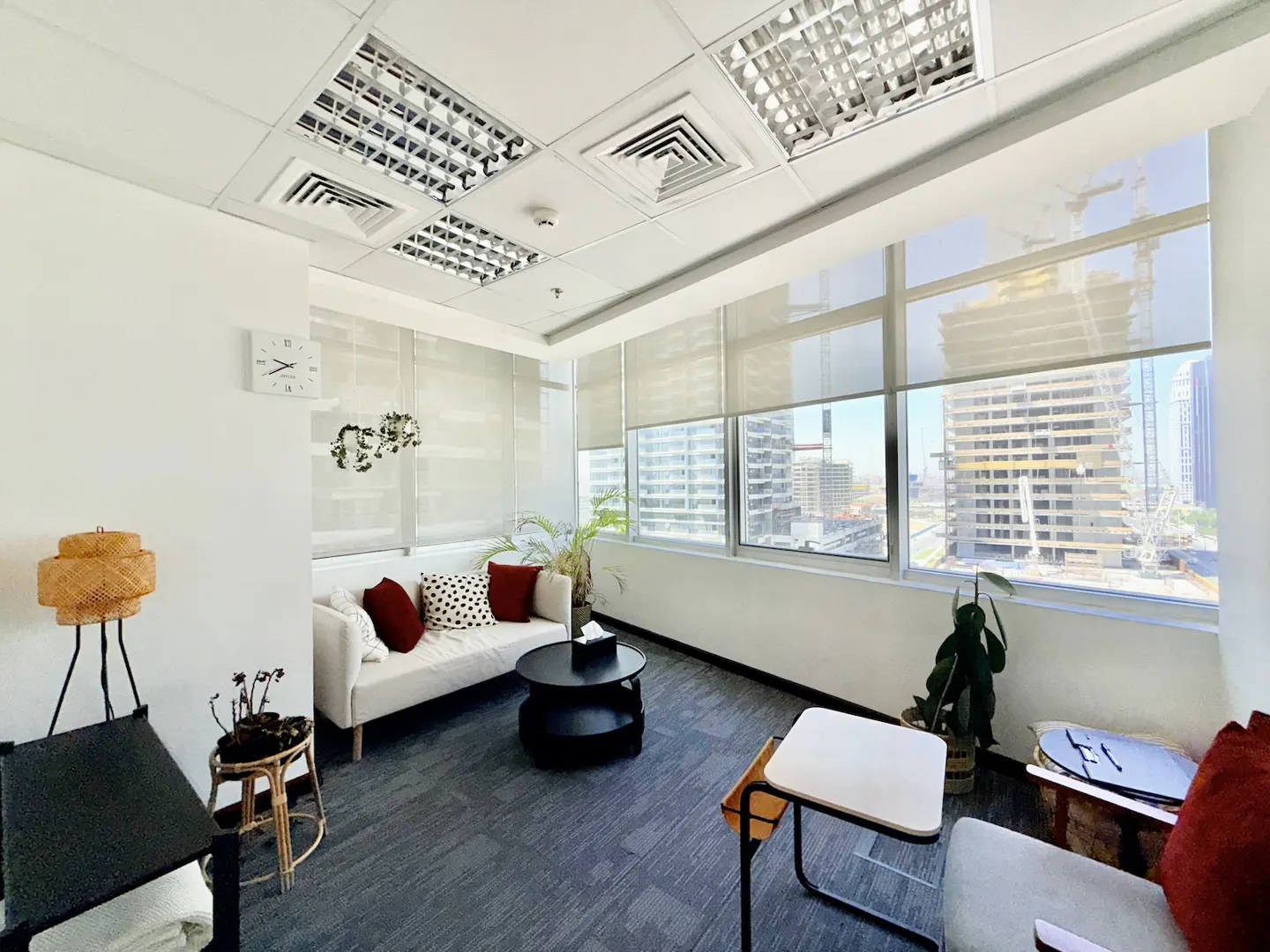 Modern office lounge with a white sofa, black coffee table, plants, and large windows overlooking a city construction site.