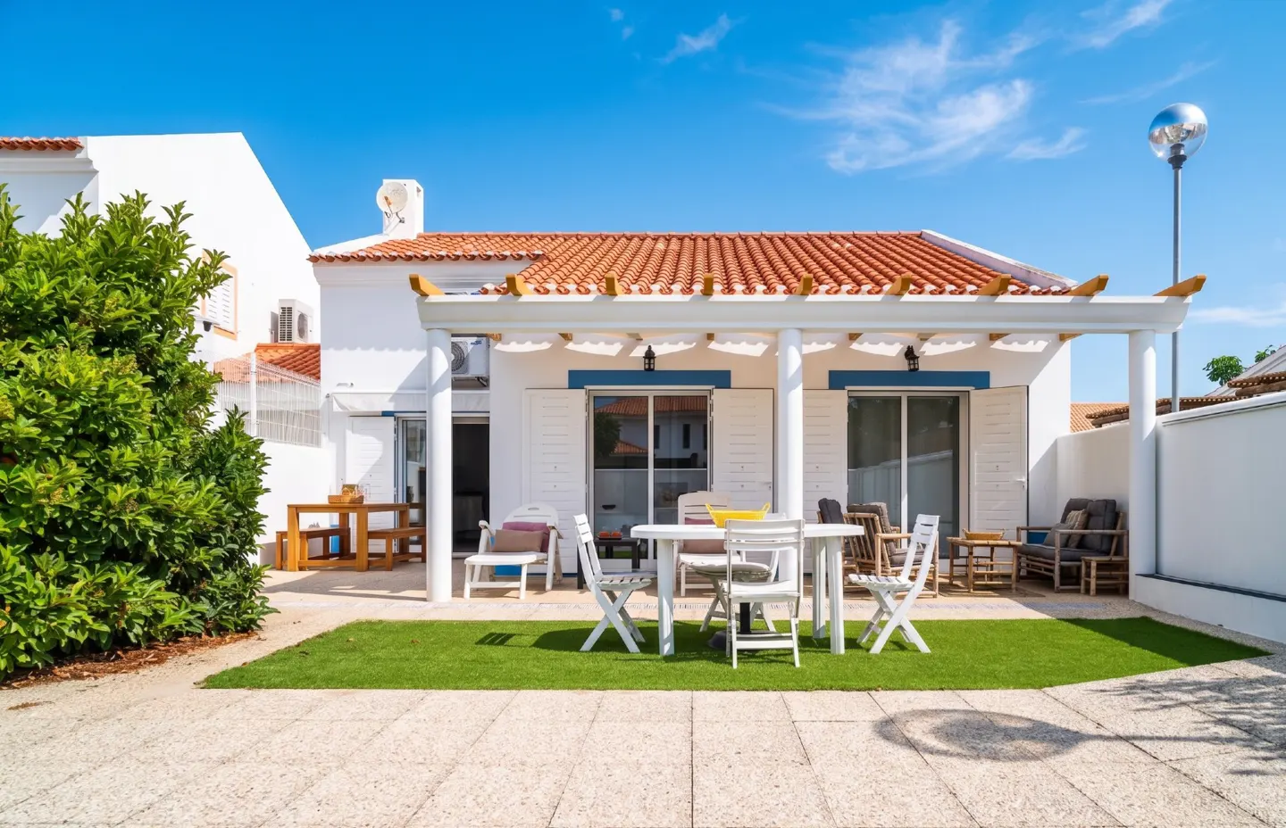 Outdoor patio with white table and chairs on green grass. White house with orange tile roof and blue sky in background.