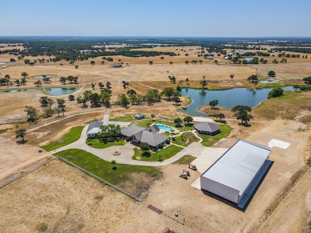 Aerial view of a rural property with a house, pool, pond, and large metal building under a clear blue sky.