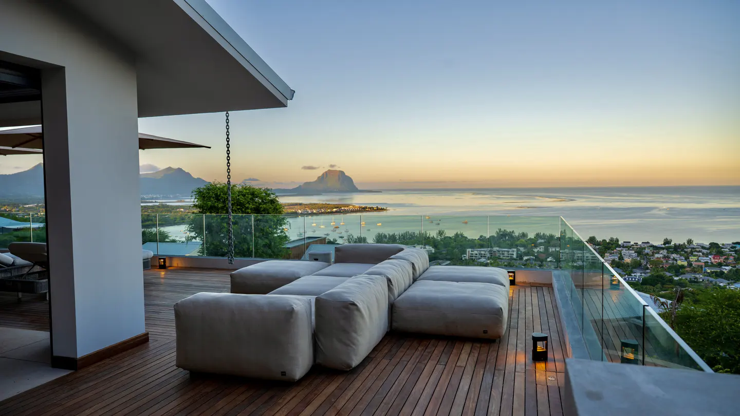 Outdoor patio with modular sofa, wood deck, and glass railing overlooking ocean and mountains at sunset.
