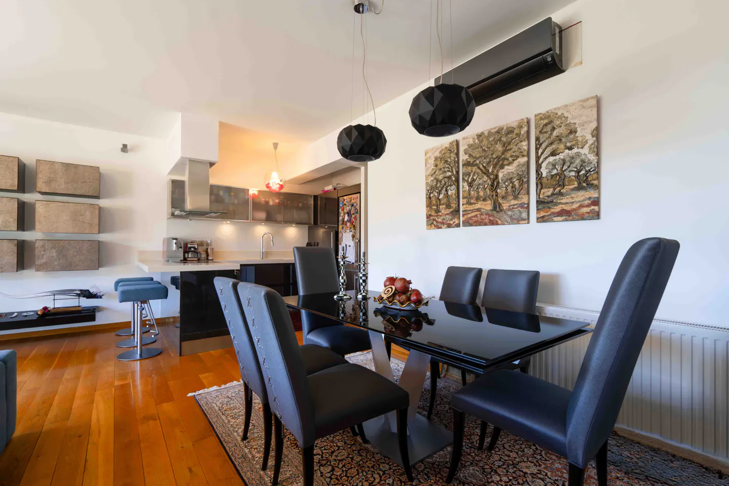 A modern dining area with a black table, gray chairs, and wooden floors. The kitchen is visible in the background.