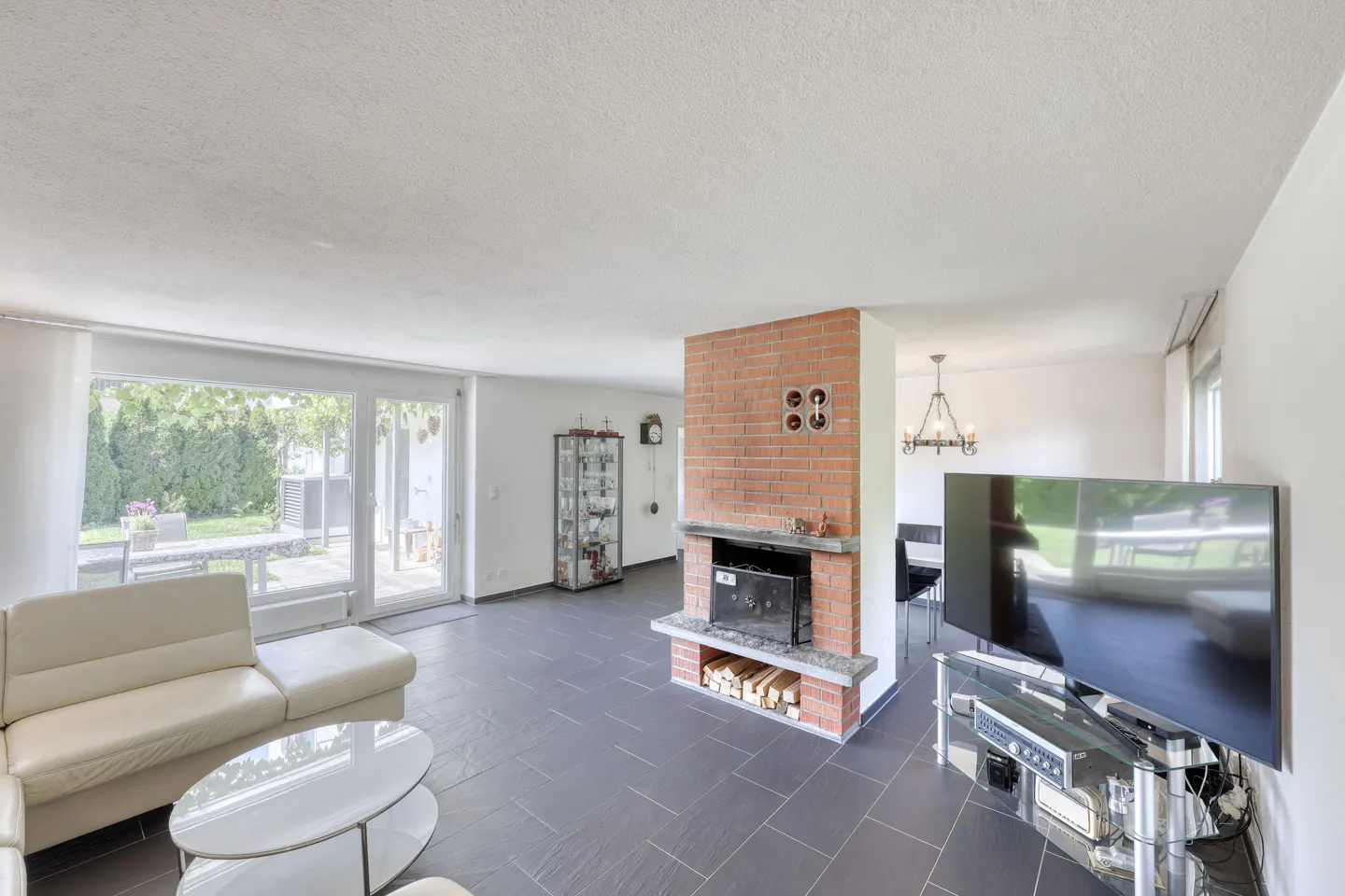 Spacious living room with white sofa, glass coffee table, brick fireplace, and large windows overlooking a patio.
