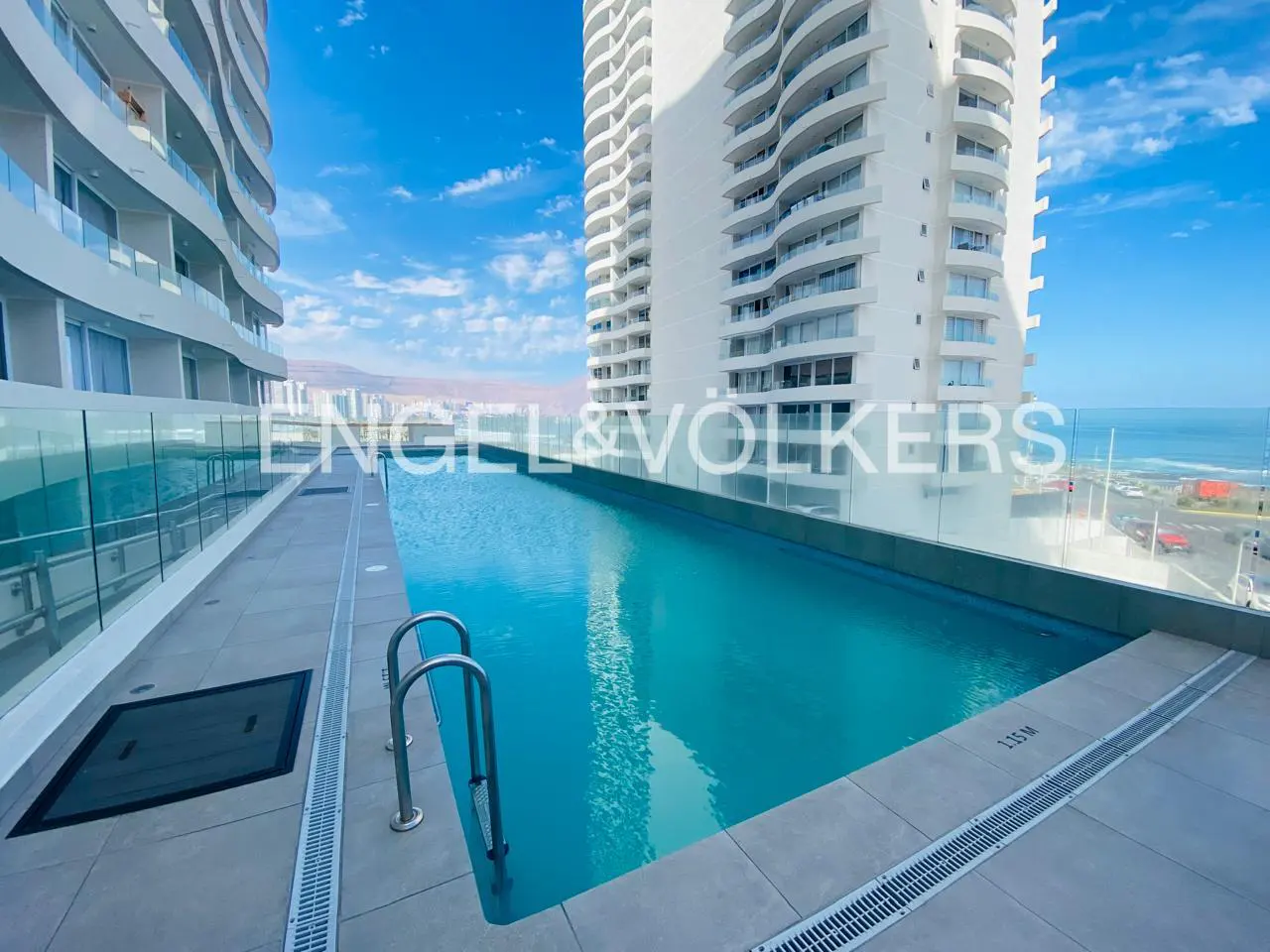 Rooftop pool with metal ladder between two white high-rise buildings. Ocean view in the background. Engel & Völkers logo on the glass.
