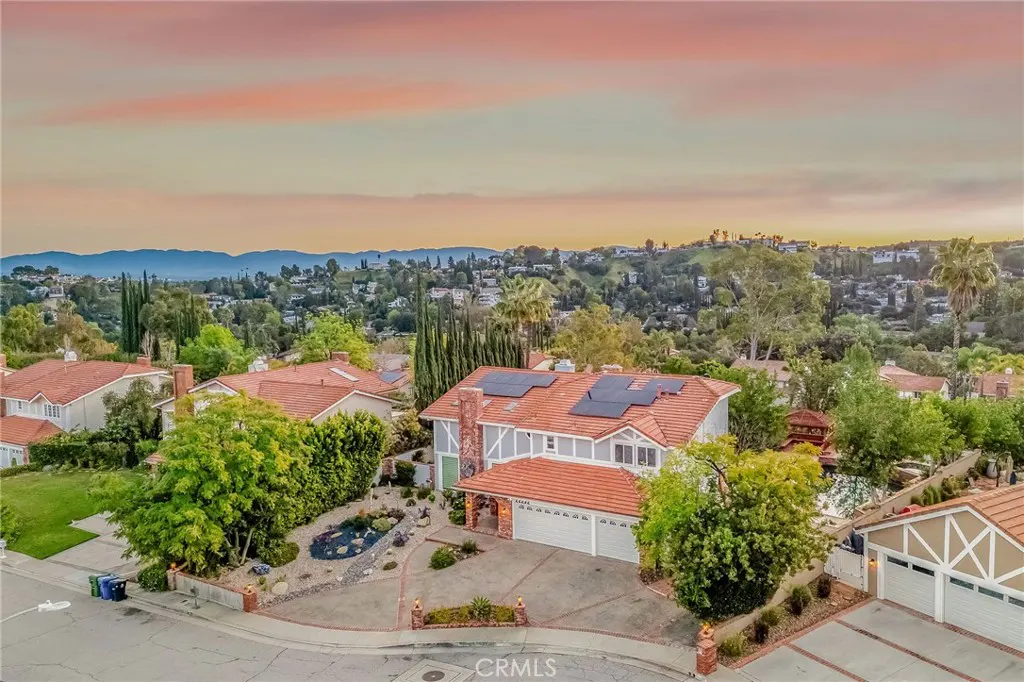 Aerial view of a two-story gray house with a red tile roof and solar panels, set in a green, hilly neighborhood at sunset.