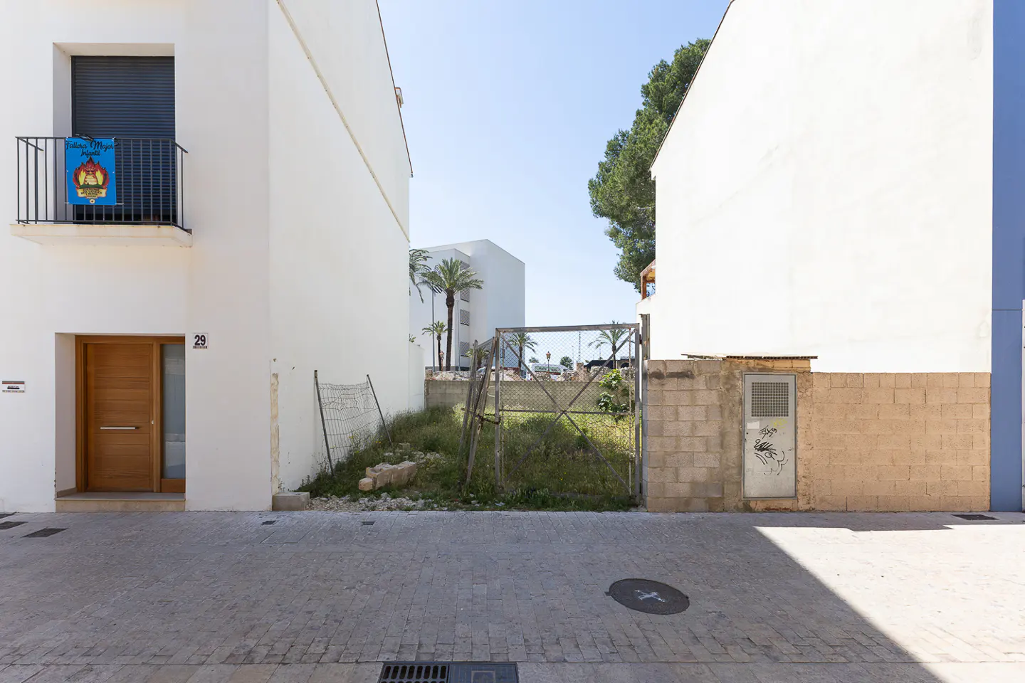 A street view shows white buildings flanking a vacant lot with a rusty gate and overgrown grass. Palm trees are visible in the background.