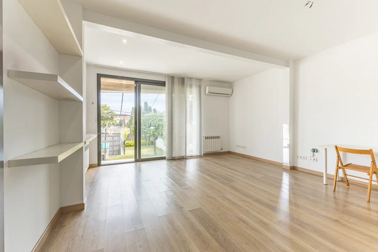 Bright, empty room with wood floors, white walls, and built-in shelves. Sliding glass doors lead to a green yard. A small desk and chair are on the right.