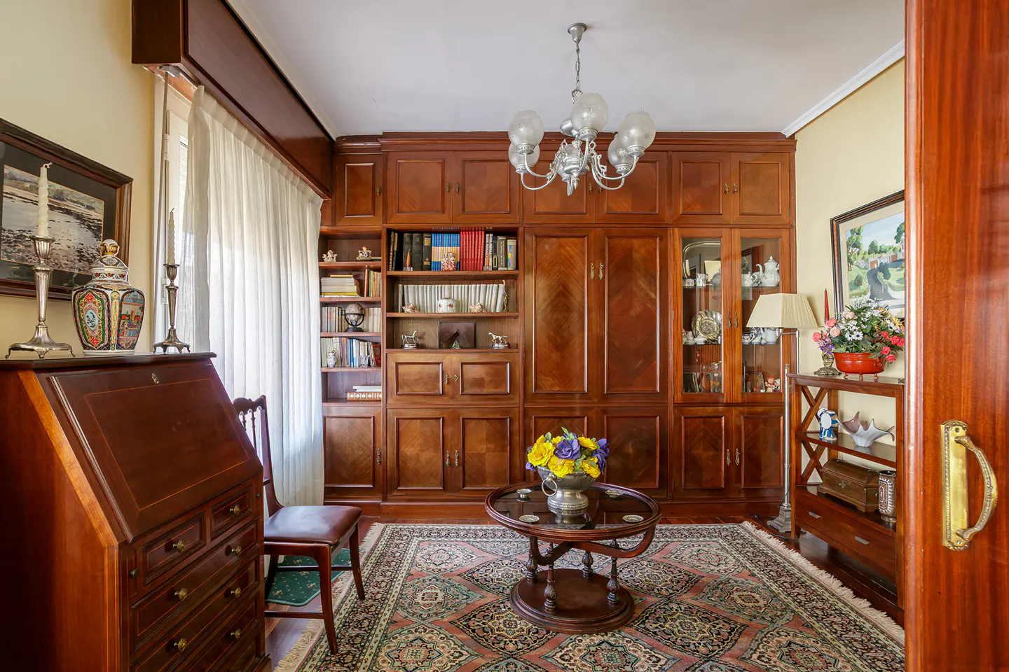 A living room with dark wood furniture, a desk, a bookcase, and a round table with flowers.