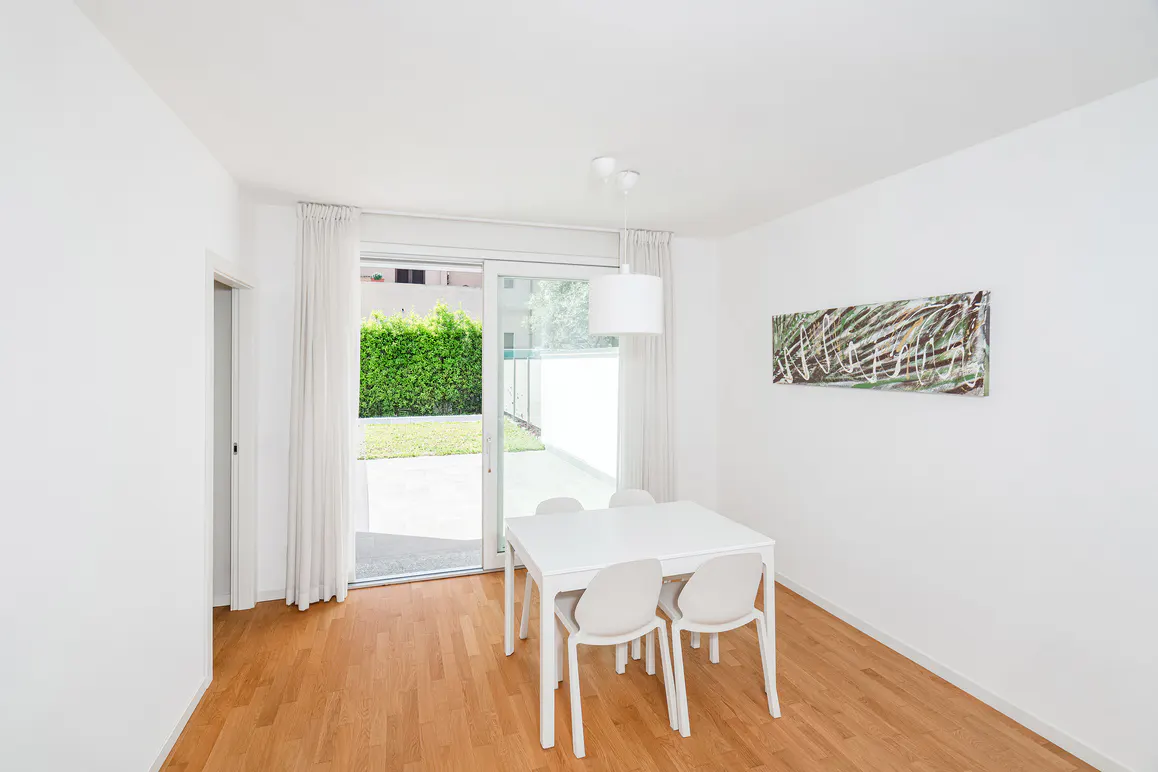 Bright dining room with white walls, wood floors, and a white table with four chairs. Sliding glass doors lead to a patio and green hedge.