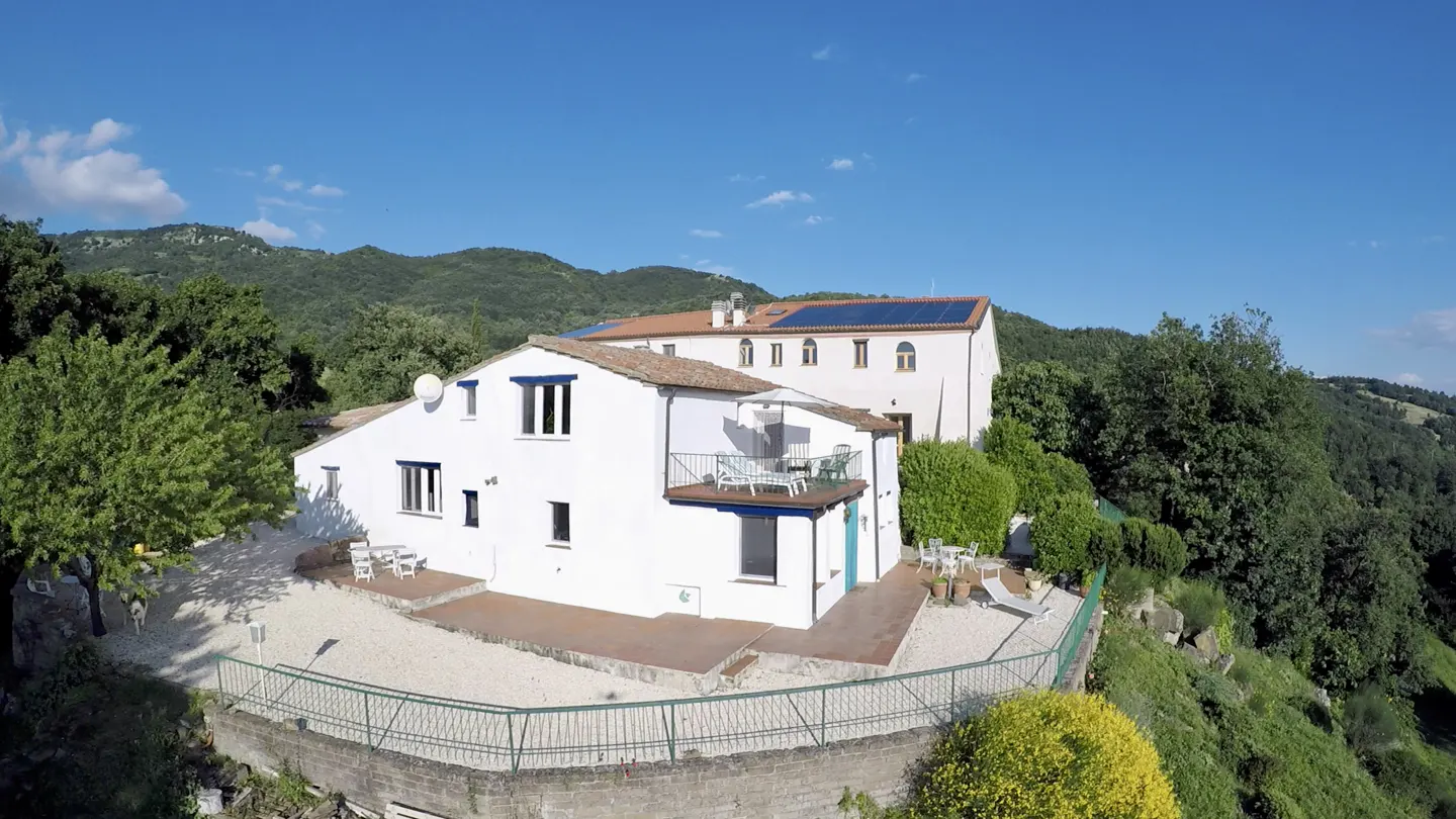 Aerial view of a white villa with a terracotta roof, balcony, and patio, set against a backdrop of lush green mountains.