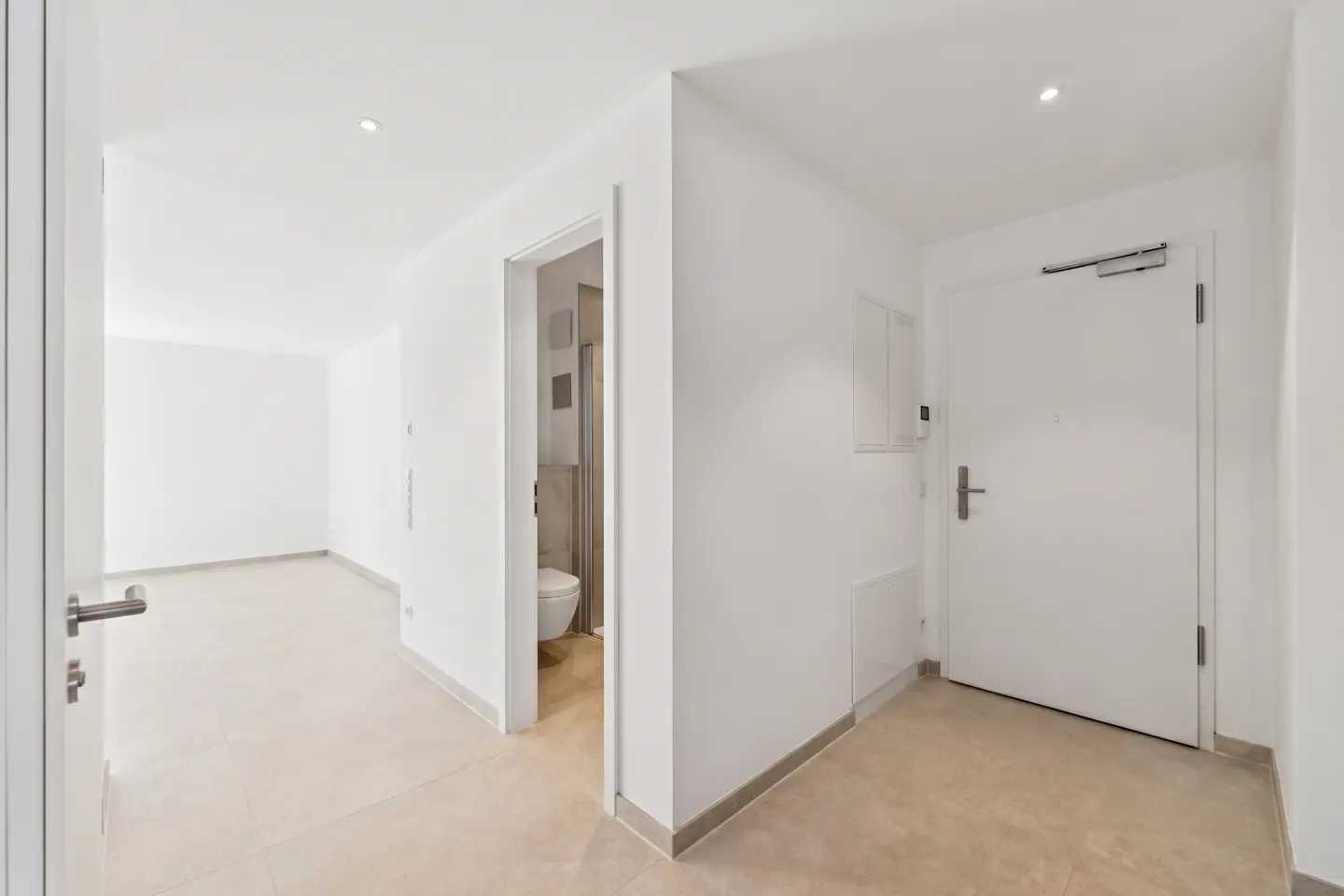 Bright, modern apartment hallway with white walls, beige tile floor, and a white front door. A bathroom is visible through an open doorway.