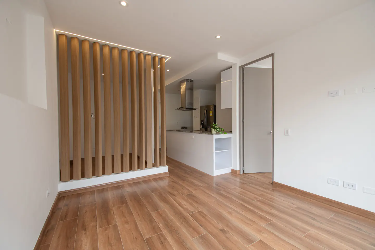 A bright, modern apartment interior with wood floors, a wood slat wall, and a glimpse of the kitchen.