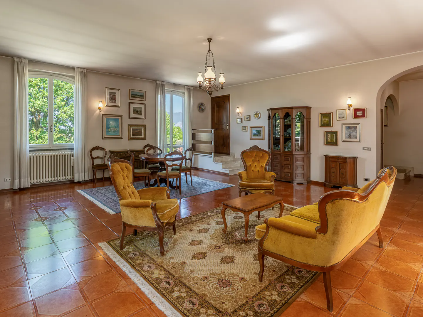 A living room with yellow velvet chairs, a wooden table, and a patterned rug on a terracotta tile floor. Windows offer a view of greenery.