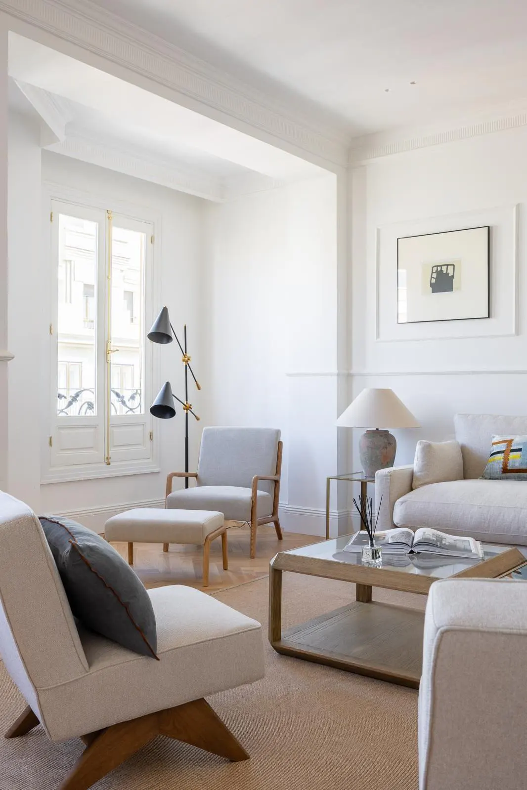 Bright living room with white walls, beige carpet, and light-colored furniture. A window lets in natural light. A modern lamp stands near a chair.