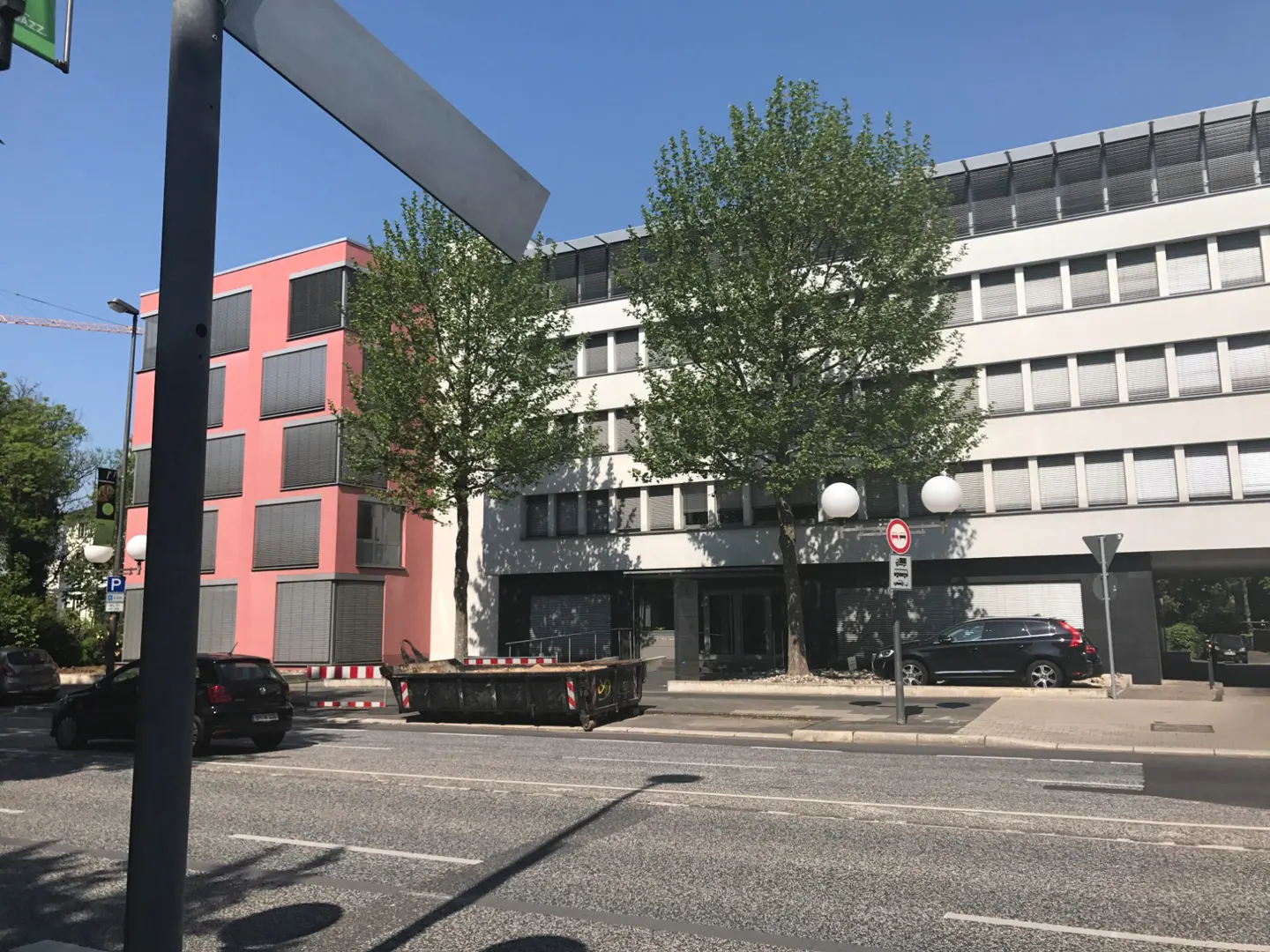 Street view of a modern building with a pink facade and a white building with many windows. A dumpster sits on the street.