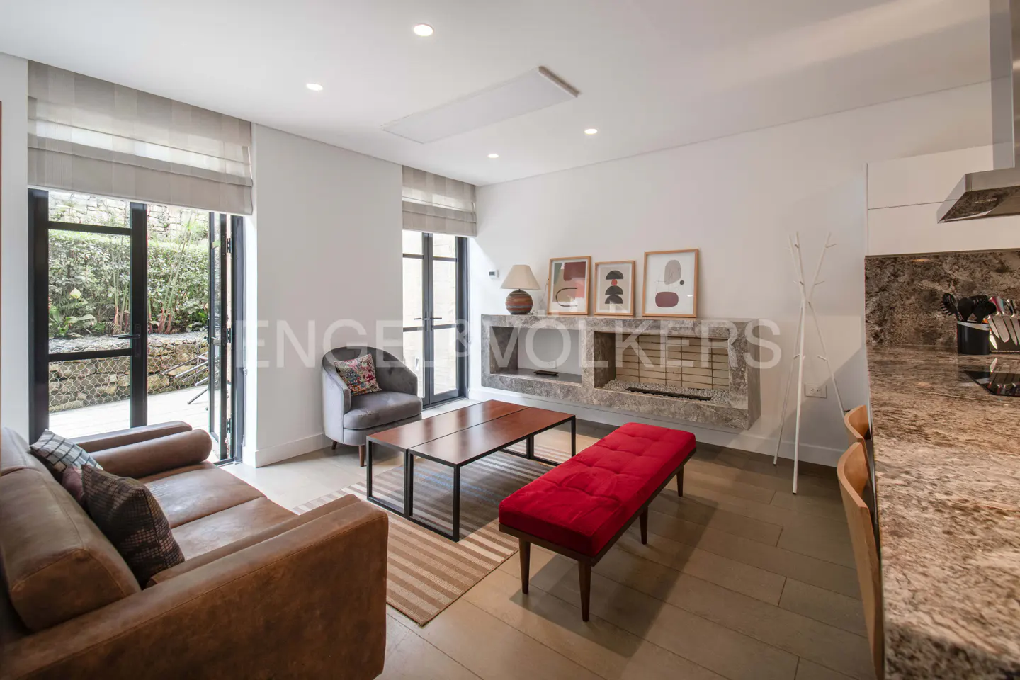 Living room with a brown leather sofa, gray armchair, red bench, and stone fireplace. Large windows overlook a green garden.