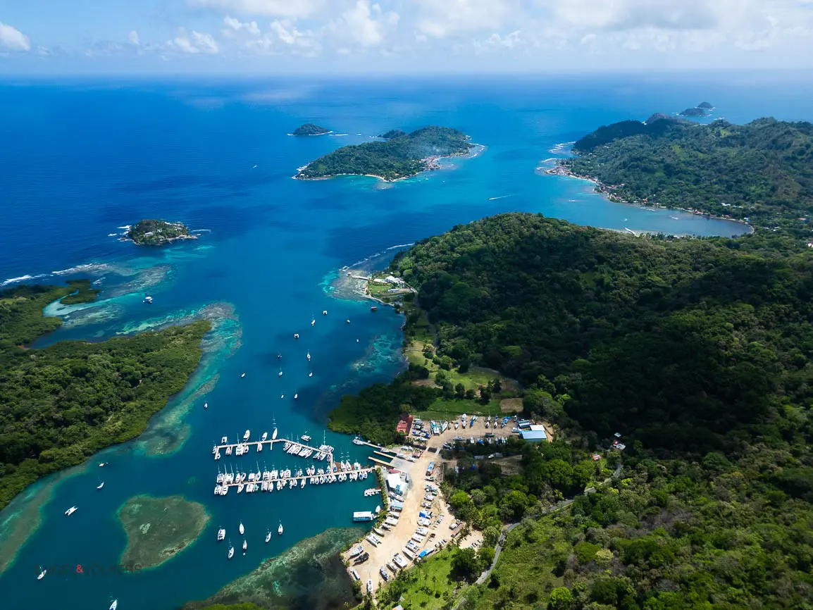 Aerial view of a marina with many boats docked, surrounded by lush green islands and blue ocean under a partly cloudy sky.