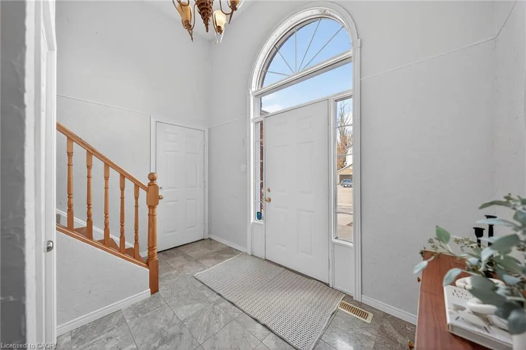 Bright foyer with white walls, gray tile floor, and a white front door with a large arched window above. A wooden staircase is to the left.