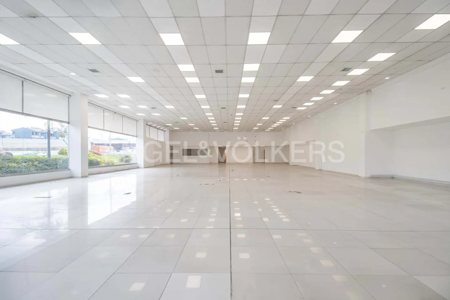 Bright, empty commercial space with white tile floors, white walls, and large windows. Overhead, a grid of fluorescent lights illuminates the room.