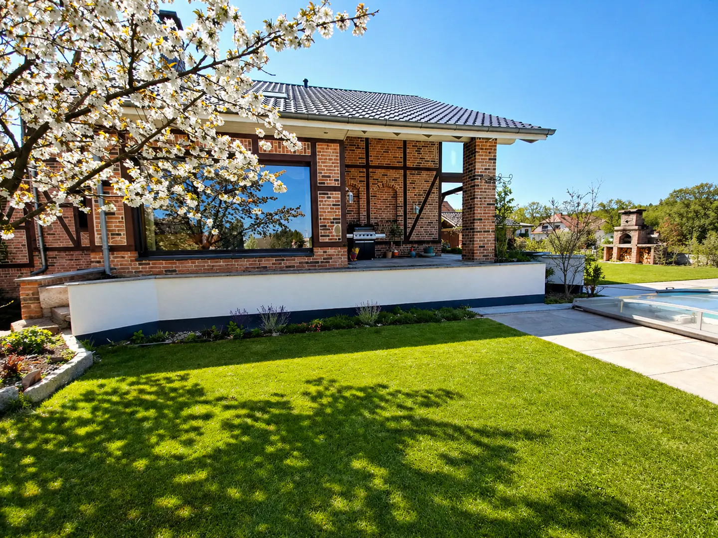 Brick house with a green lawn and blooming tree. A pool and outdoor fireplace are in the background.