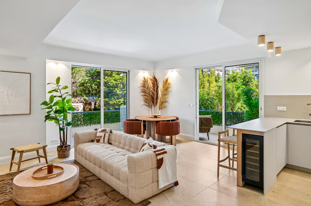 Bright, modern living room with a beige sofa, round coffee table, and dining area with brown chairs. Sliding glass doors open to a green outdoor space.