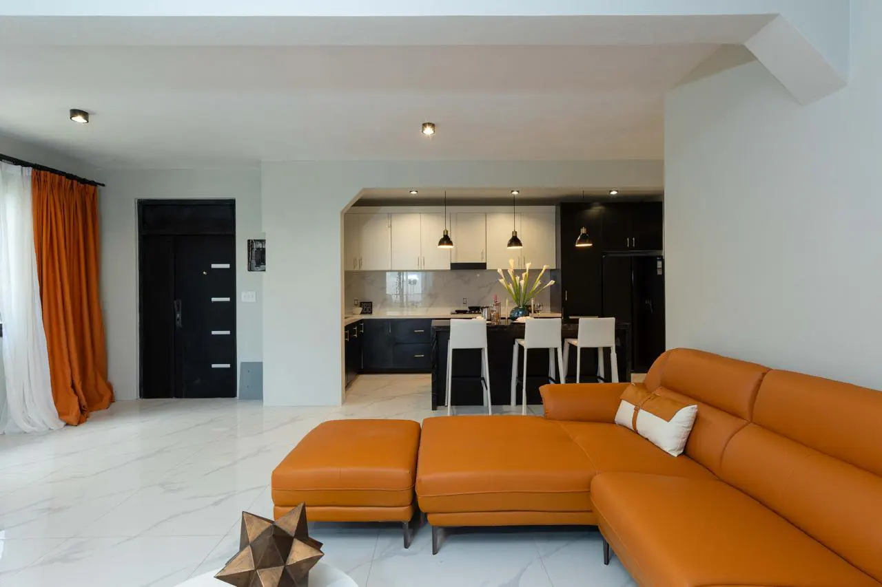 Open concept living space with orange leather sectional sofa, white marble floors, and black and white kitchen.