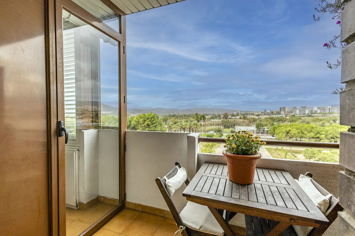 Balcony view with a wooden table, chairs, and potted yellow flowers. A door opens to a scenic landscape of trees and buildings under a blue sky.