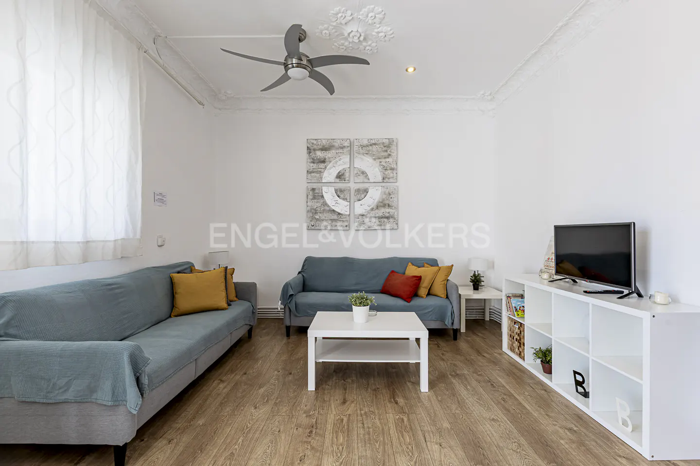 Bright living room with wood floors, white walls, and two blue sofas with yellow and red pillows. A white table and TV stand are also present.
