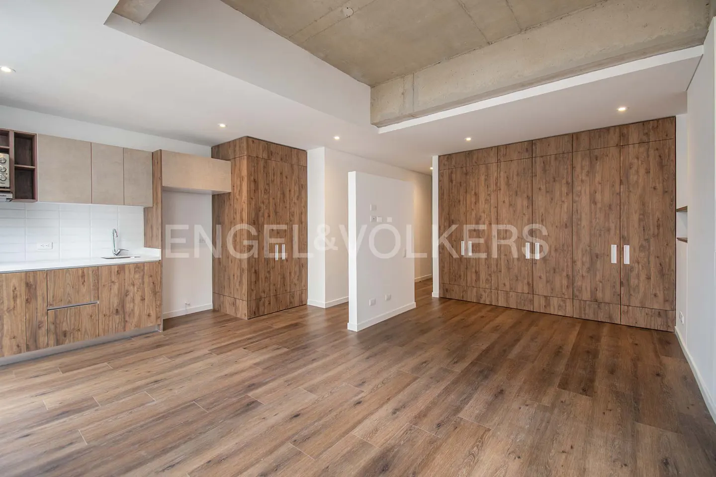 A modern apartment interior with wood floors, cabinets, and white walls. The kitchen area is visible on the left.