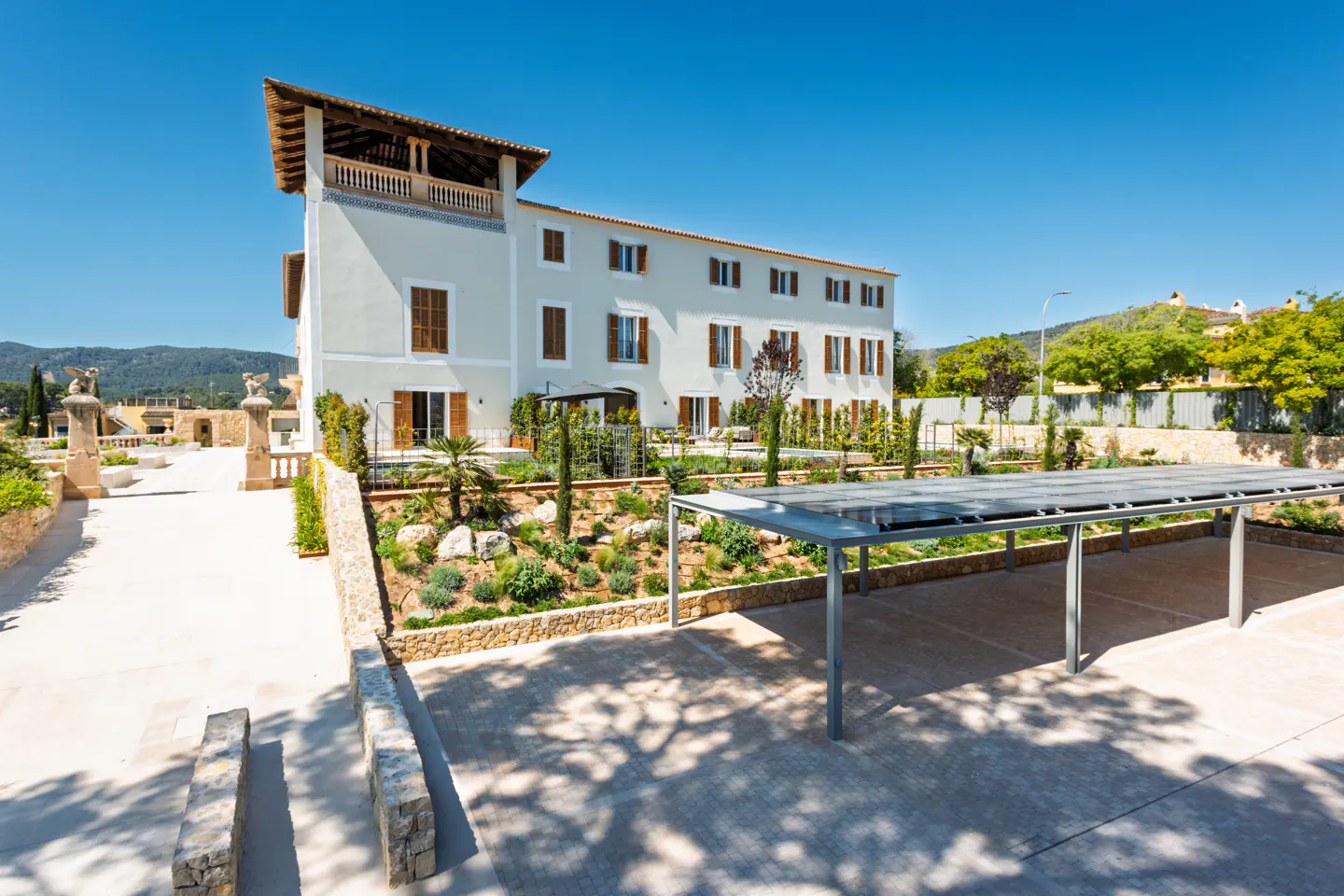Exterior view of a large, light-gray building with brown shutters, a garden, and a covered parking area under a clear blue sky.
