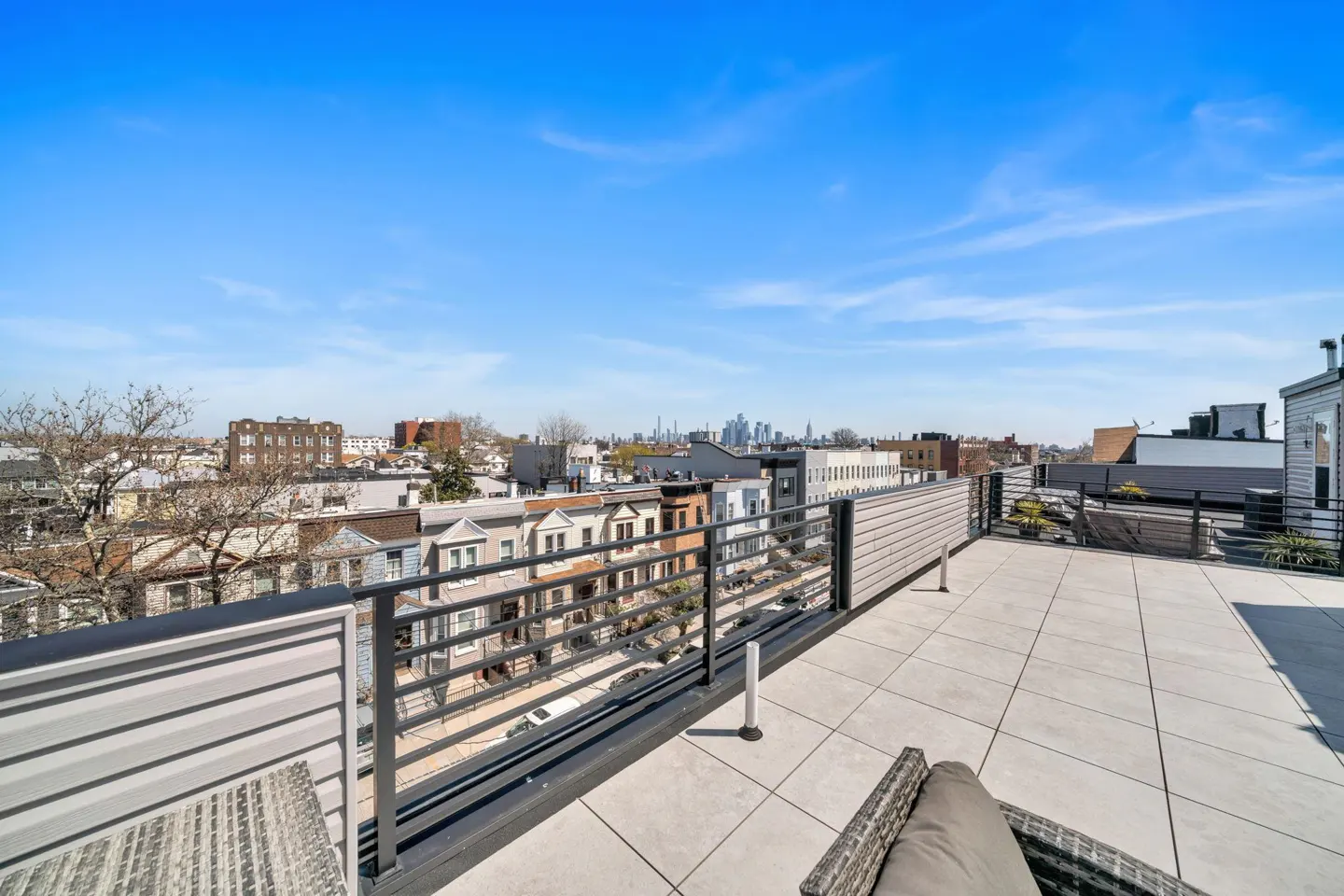 Rooftop patio with gray tiles, metal railing, and wicker furniture. City skyline visible in the background under a blue sky.