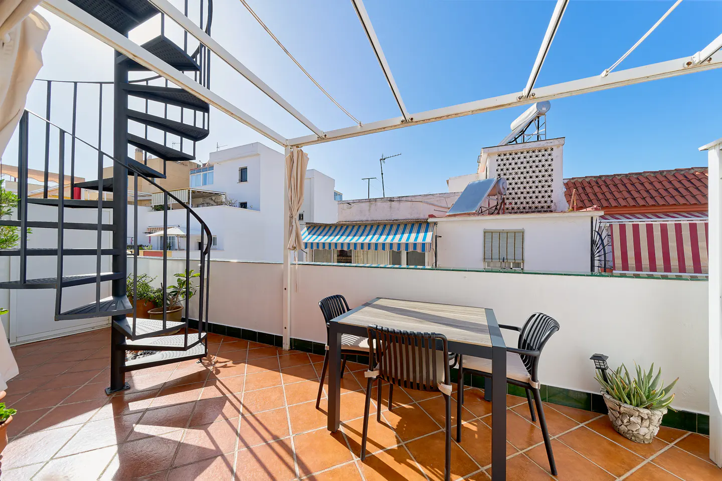 Outdoor patio with a black spiral staircase, table, and chairs on terracotta tile. White buildings and a blue sky are in the background.