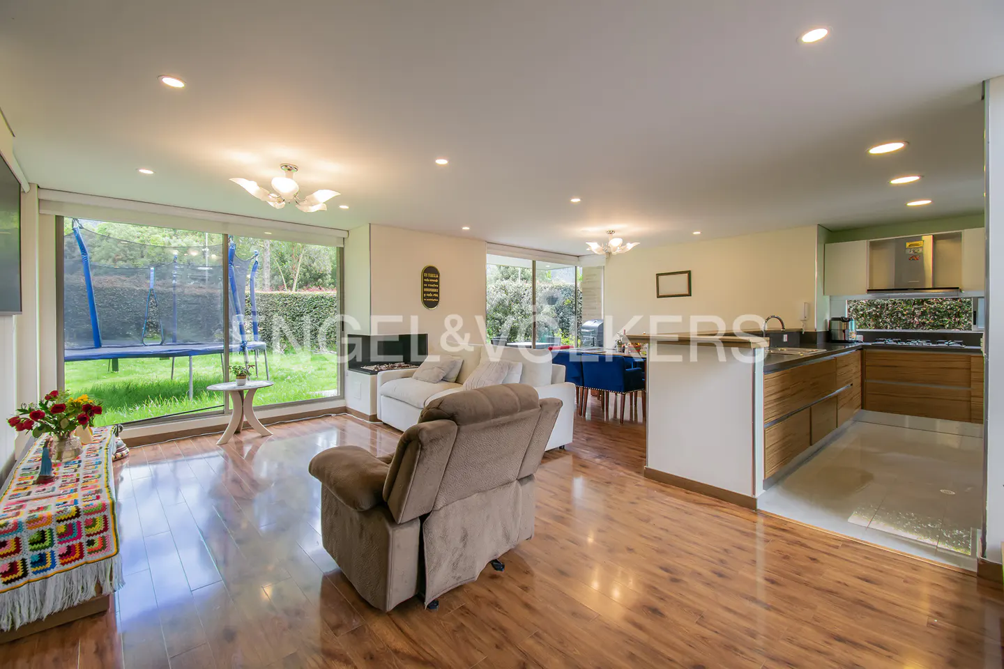 Bright, open living space with wood floors, a brown recliner, and a white sofa. A trampoline is visible through the large windows.