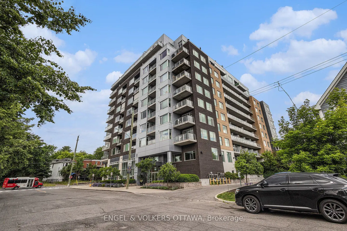 Exterior view of a modern, multi-story condo building with balconies, a black car parked in front, and green trees surrounding it.