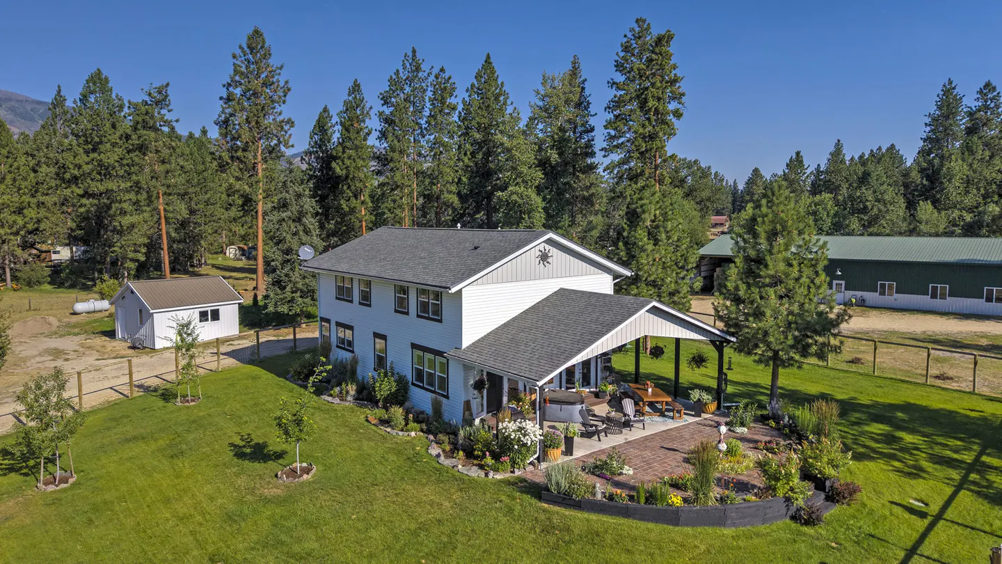 Aerial view of a two-story white house with a gray roof, surrounded by green lawn, trees, and a brick patio with outdoor furniture.