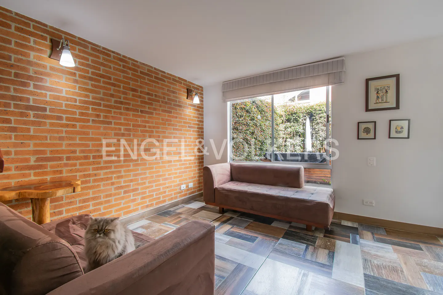 Living room with a brick wall, brown sofas, and a gray cat sitting on one of them. A window overlooks a garden with a fountain.
