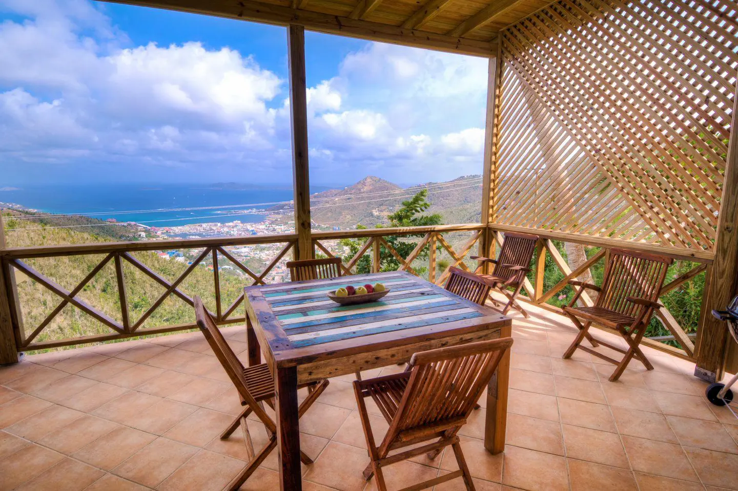 Covered patio with a wooden table, chairs, and a view of the ocean and city below.