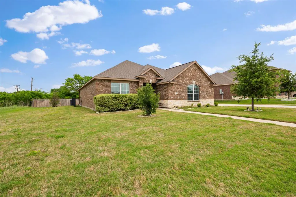 A single-story brick house with a brown roof sits on a green lawn under a blue sky with white clouds.