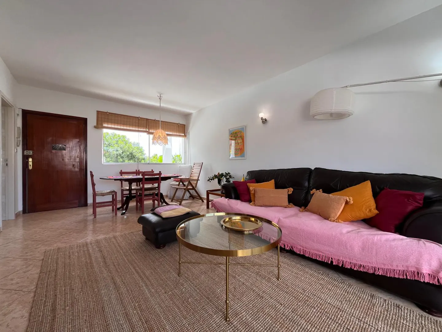 Living room with a brown leather sofa, pink throw, and colorful pillows. A round gold coffee table sits on a jute rug. A dining table and chairs are near a window.