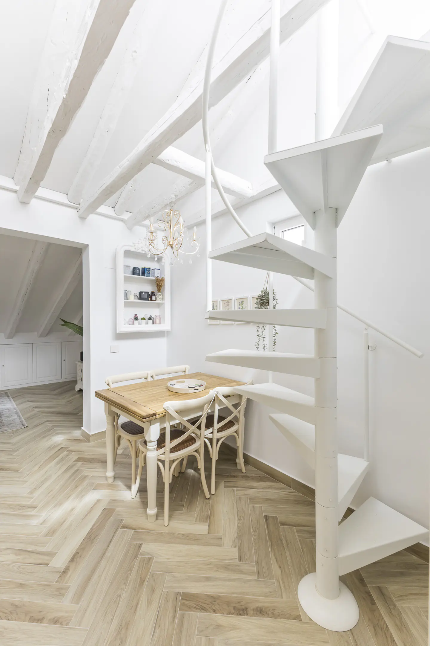 Bright, modern dining area with a wooden table, white chairs, and a white spiral staircase. Herringbone wood floors and white beamed ceilings add texture.
