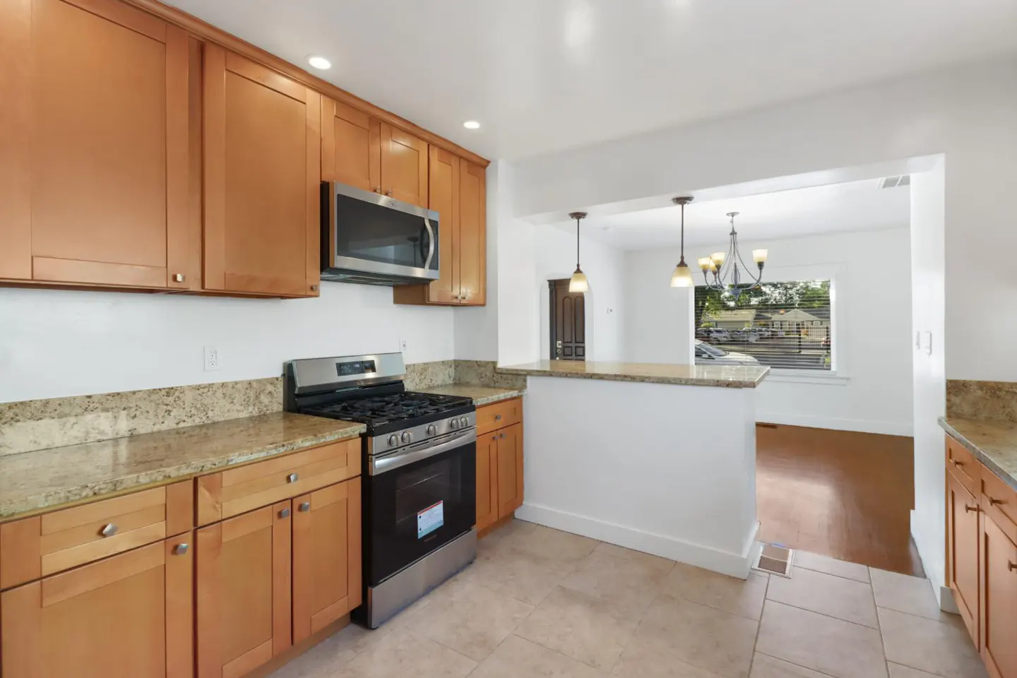 Bright kitchen with tan cabinets, granite countertops, and stainless steel appliances. View into dining area with wood floors.