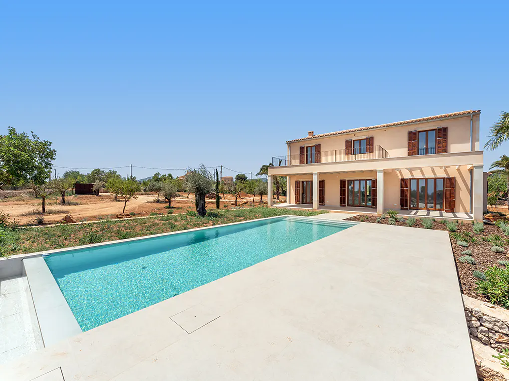 Two-story beige house with brown shutters, a pool, and a clear blue sky.