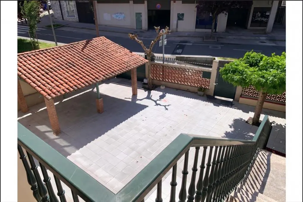 View from stairs of a patio with a red-tiled roof structure, gray pavers, a brick fence, and trees, with a street in the background.