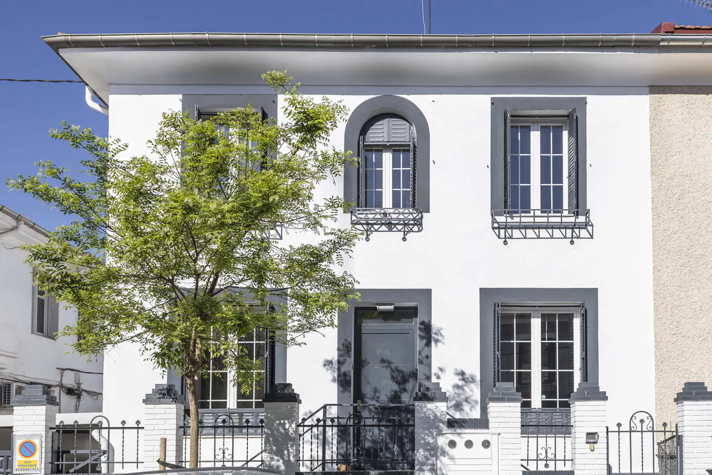 Two-story white house with gray trim, black iron fence, and a green tree in front. Blue sky in the background.