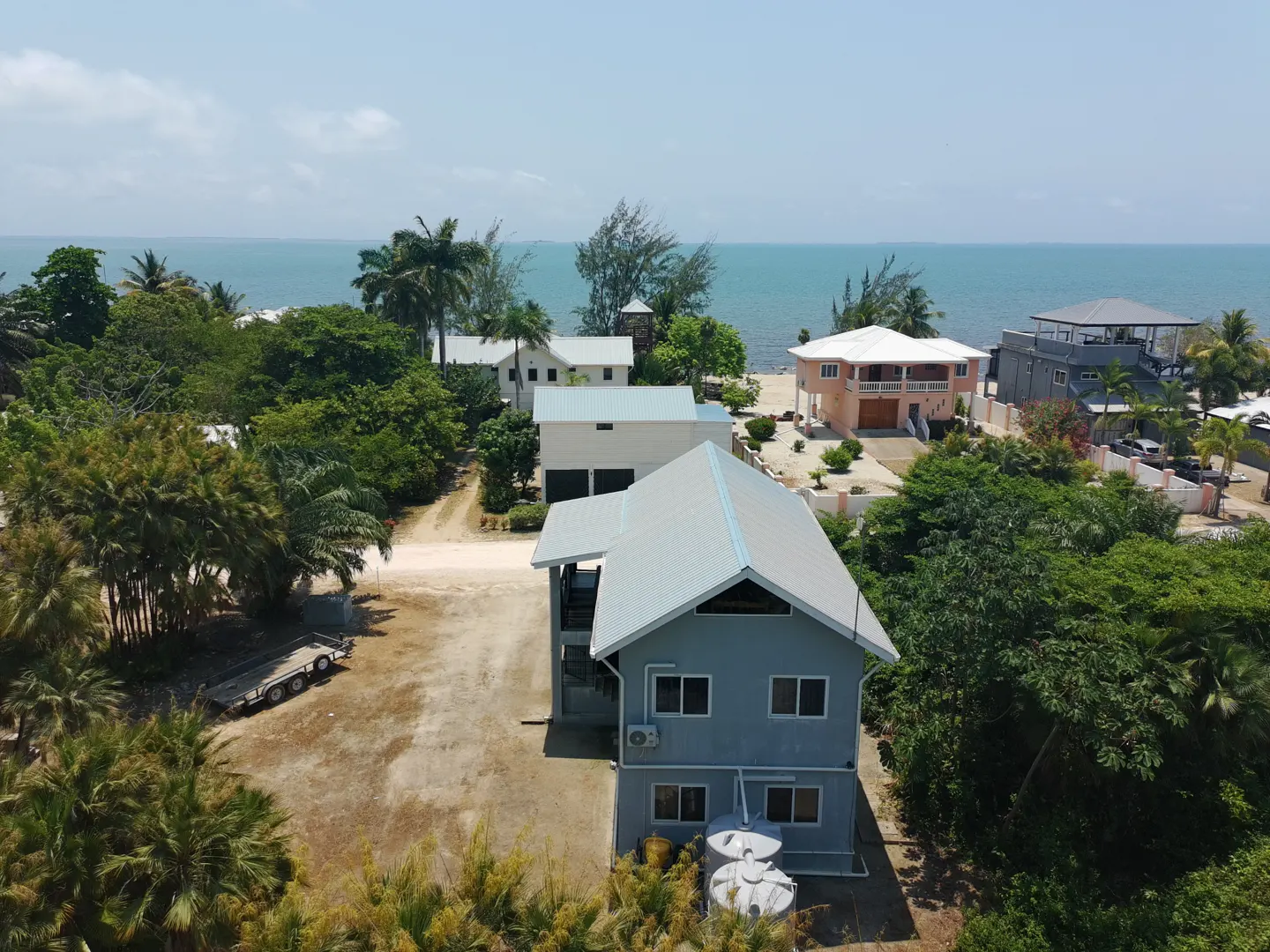 Aerial view of a two-story blue house with a silver roof near the ocean, surrounded by lush green trees and other houses.